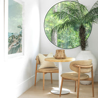 Round bistro table with a timber top and white pedestal base, paired with leather dining chairs in a cozy breakfast nook, accented by a large round window and a white earthenware vase with lush greenery