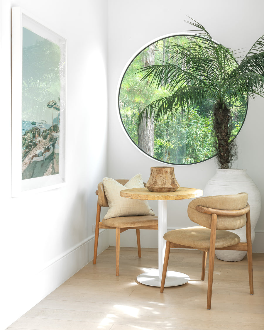 Round bistro table with a timber top and white pedestal base, paired with leather dining chairs in a cozy breakfast nook, accented by a large round window and a white earthenware vase with lush greenery