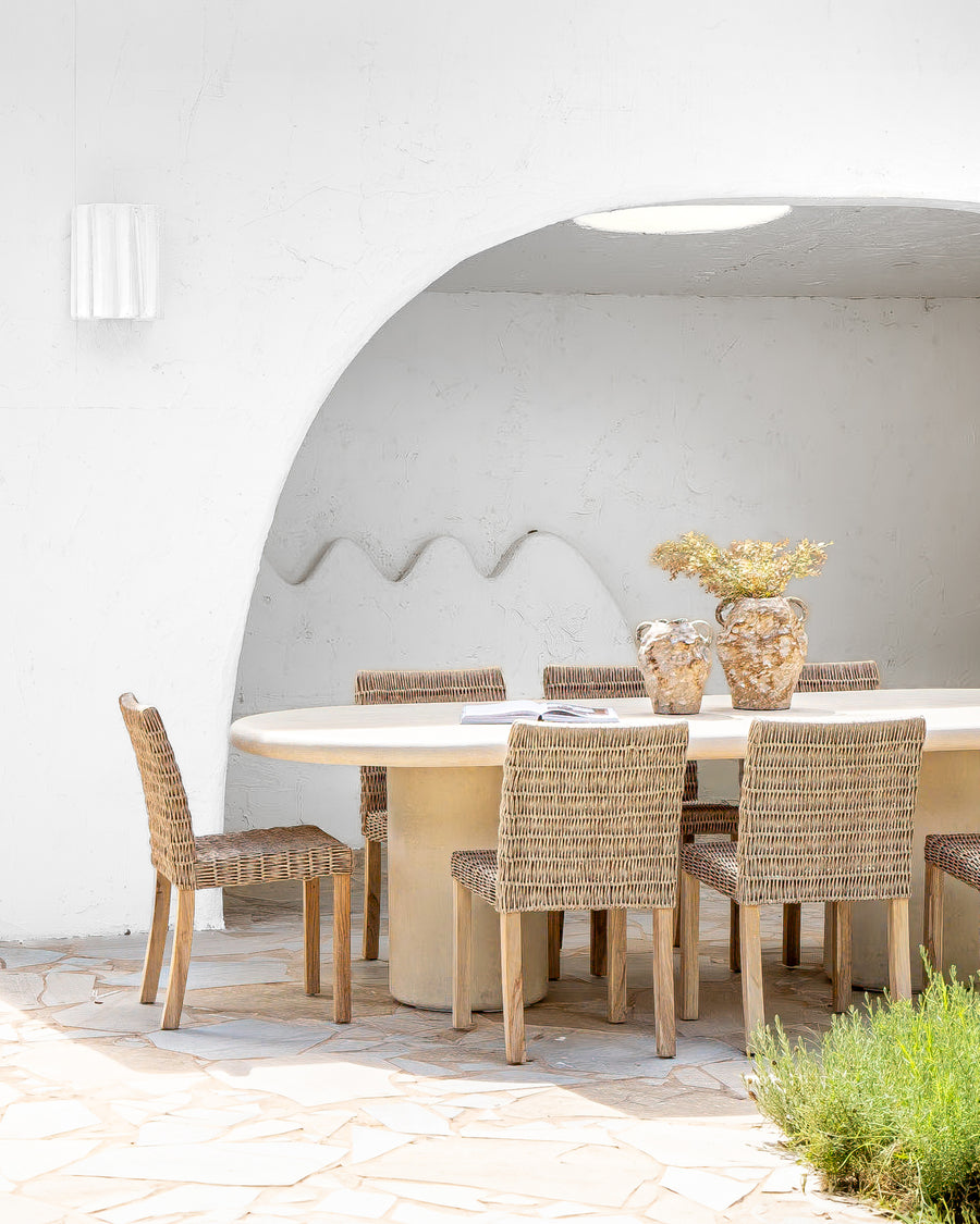 Round concrete dining table paired with woven dining chairs, styled under a white archway with natural vases and dried foliage, creating a tranquil alfresco dining space.