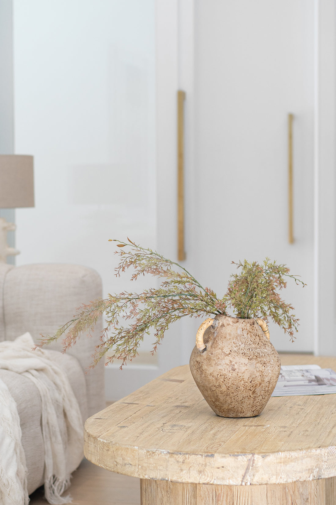 A rustic ceramic vase with woven handles, filled with dried foliage, sits atop a natural timber coffee table in a serene living space. Styled with organic textures and neutral tones, perfect for modern interiors