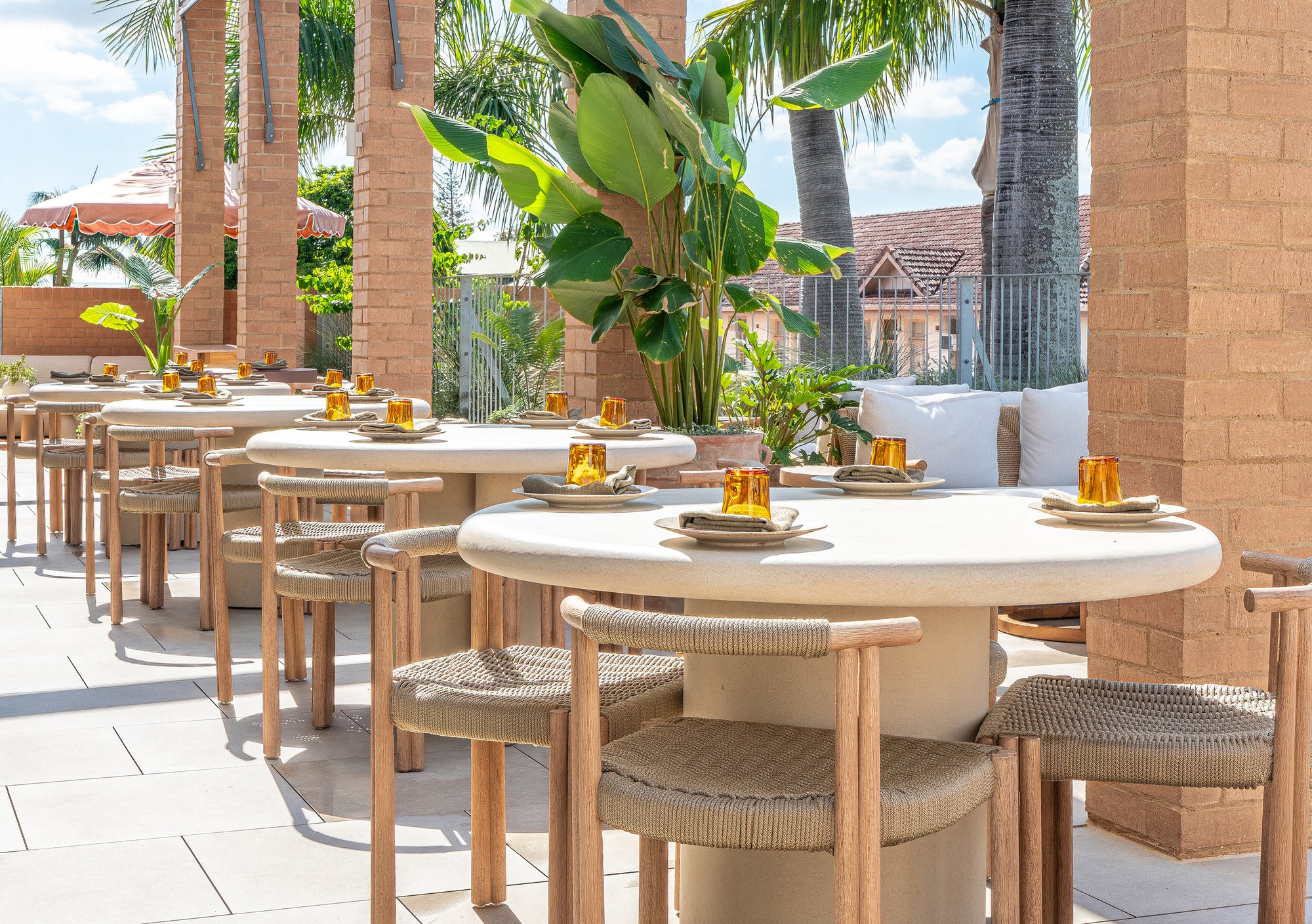 Salalah dining table arranged across a sunlit rooftop setting with woven chairs warm brick architecture and lush tropical planting creating a relaxed outdoor dining atmosphere