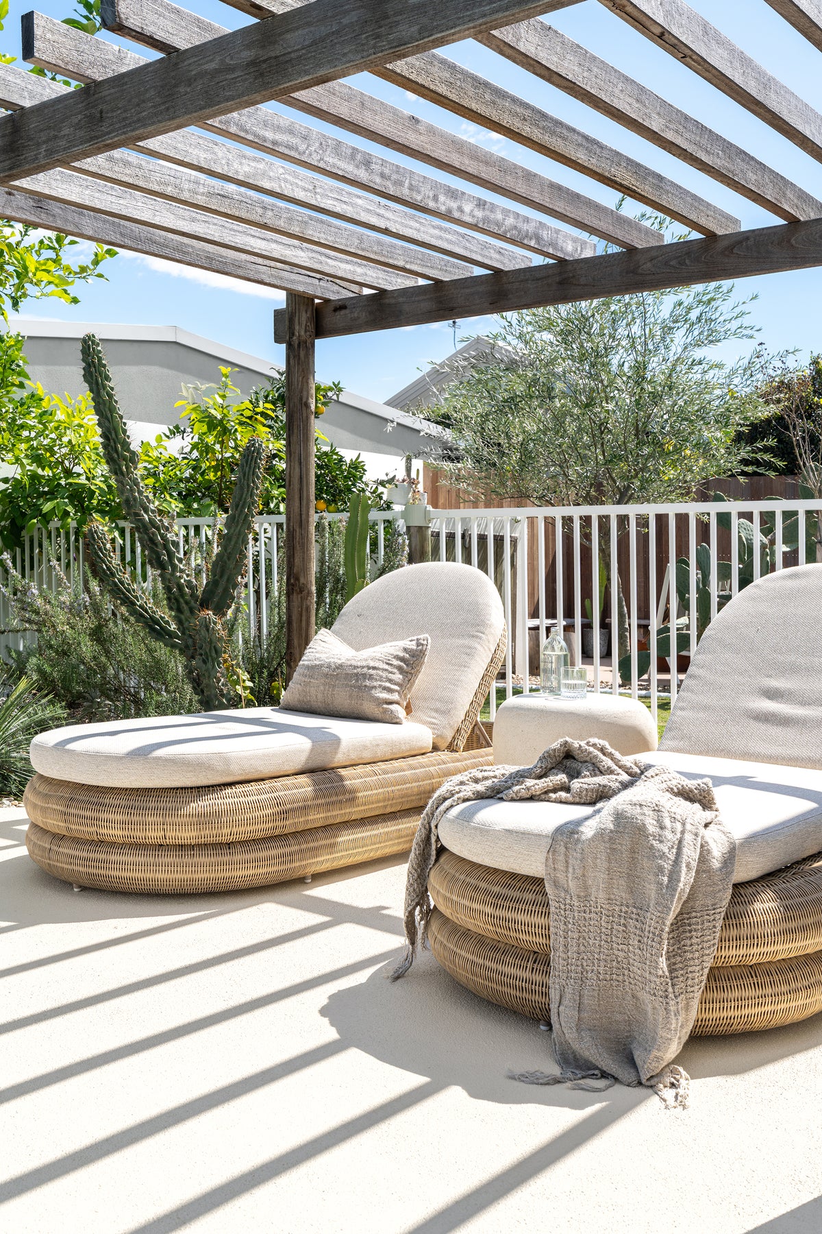 Two Satori Outdoor Sun Loungers with natural woven bases and cream cushions beneath a rustic timber pergola, surrounded by lush coastal garden greenery.