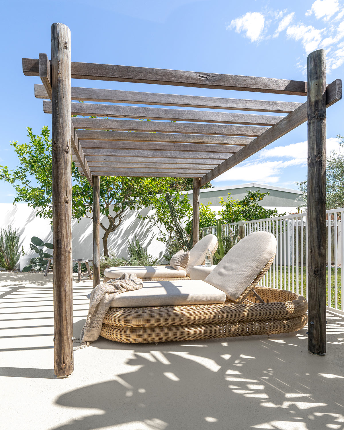 Two Satori Outdoor Sun Loungers with natural woven bases and cream cushions beneath a timber pergola in a sunlit coastal garden.
