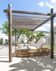 Two Satori Outdoor Sun Loungers with natural woven bases and cream cushions beneath a timber pergola in a sunlit coastal garden.