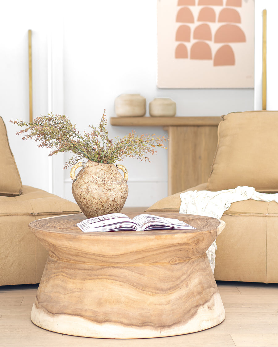 Solid timber coffee table styled with a Naga Vase filled with dried foliage, positioned between suede lounges in a warm, earthy-toned living space with organic decor elements