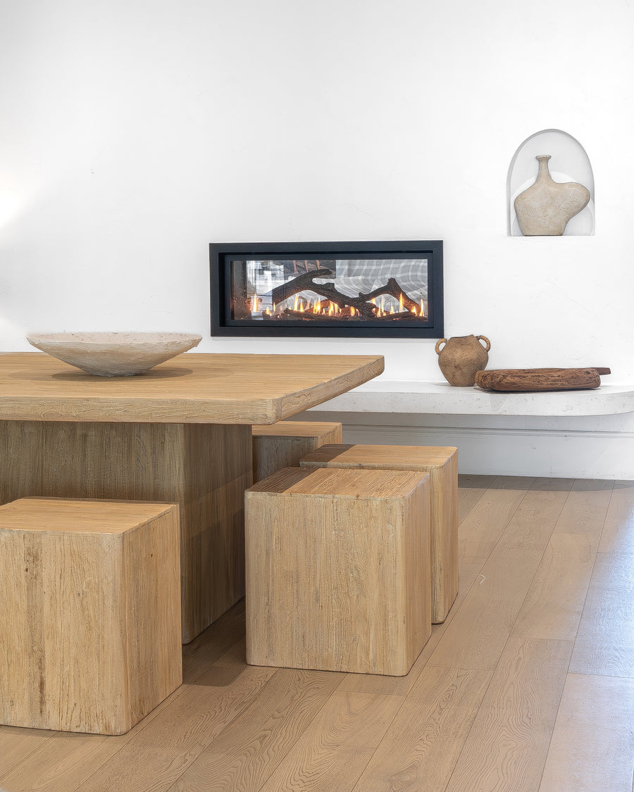 Solid timber dining table paired with matching block-style stools, styled with a textured ceramic bowl, Vase, and hand-carved wooden tray, set against a modern fireplace for a warm and inviting dining space