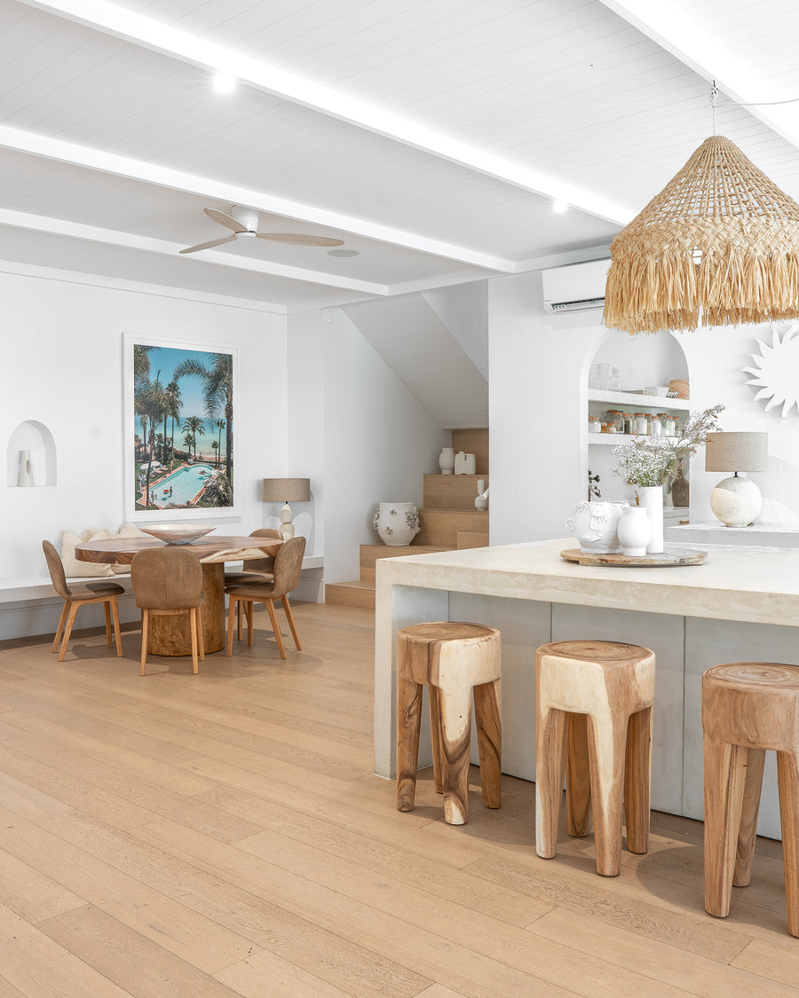 Teak bar stools paired with a stone kitchen island, complemented by a woven rattan pendant light and a round timber dining table with leather dining chairs, creating a warm and inviting modern dining space