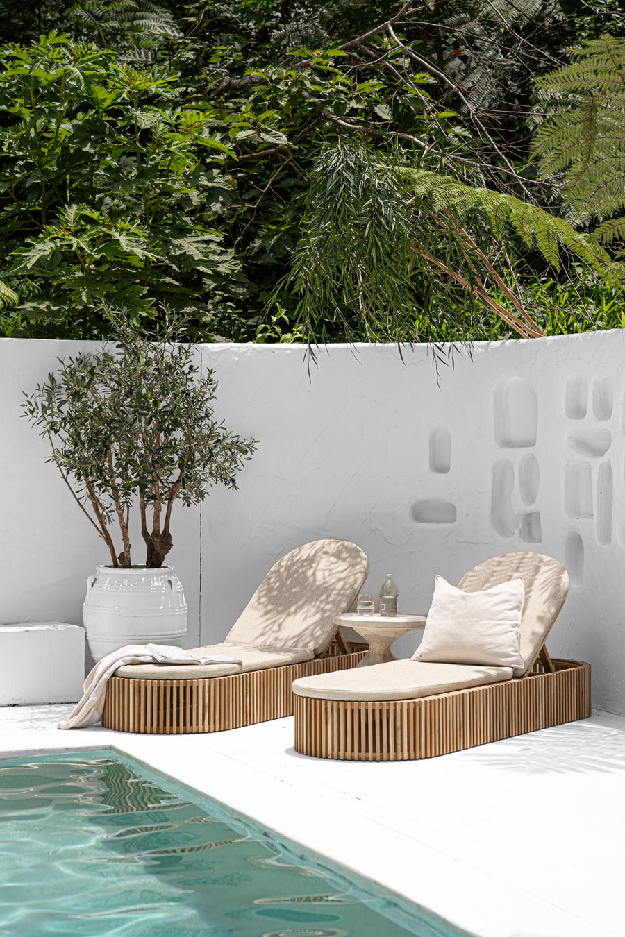 Luxury outdoor teak sun loungers with plush cushions, set against a white Mediterranean-style backdrop with lush greenery, creating a serene poolside retreat