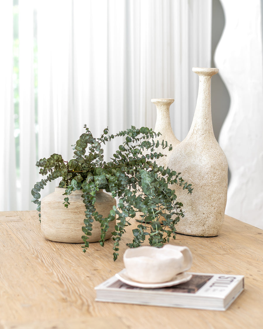 Uniqwa Collections a collection of textured ceramic vases with lush greenery arranged on a natural wooden table, complemented by a sculptural ceramic cup and a design magazine, creating a serene and organic aesthetic