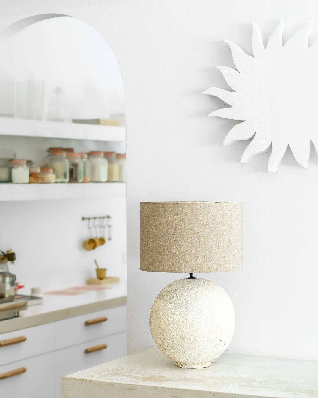 Uniqwa Collections textured table lamp with a neutral fabric shade styled on a stone console against a white wall, with an arched mirror reflecting an organic kitchen space