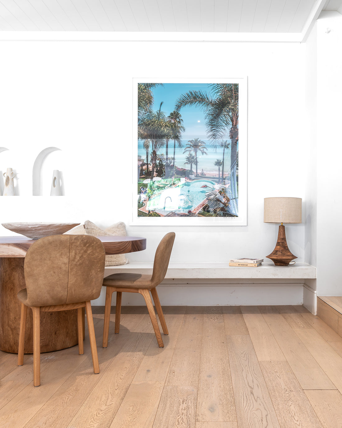 Timber dining table styled with leather dining chairs, a sculptural bowl, and textured cushions, complemented by a coastal-inspired wall art piece and a warm-toned wooden table lamp in a refined dining nook