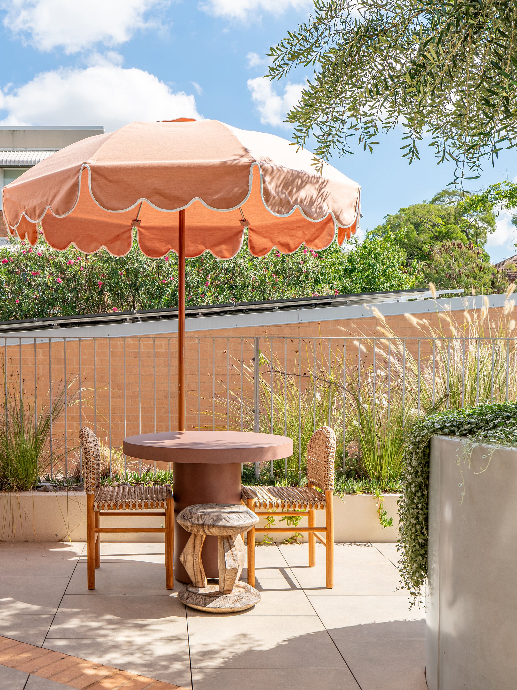 Tsuana stool styled beside a round outdoor table under a soft pink umbrella with woven chairs and lush greenery creating a relaxed rooftop setting