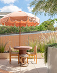 Tsuana stool styled beside a round outdoor table under a soft pink umbrella with woven chairs and lush greenery creating a relaxed rooftop setting