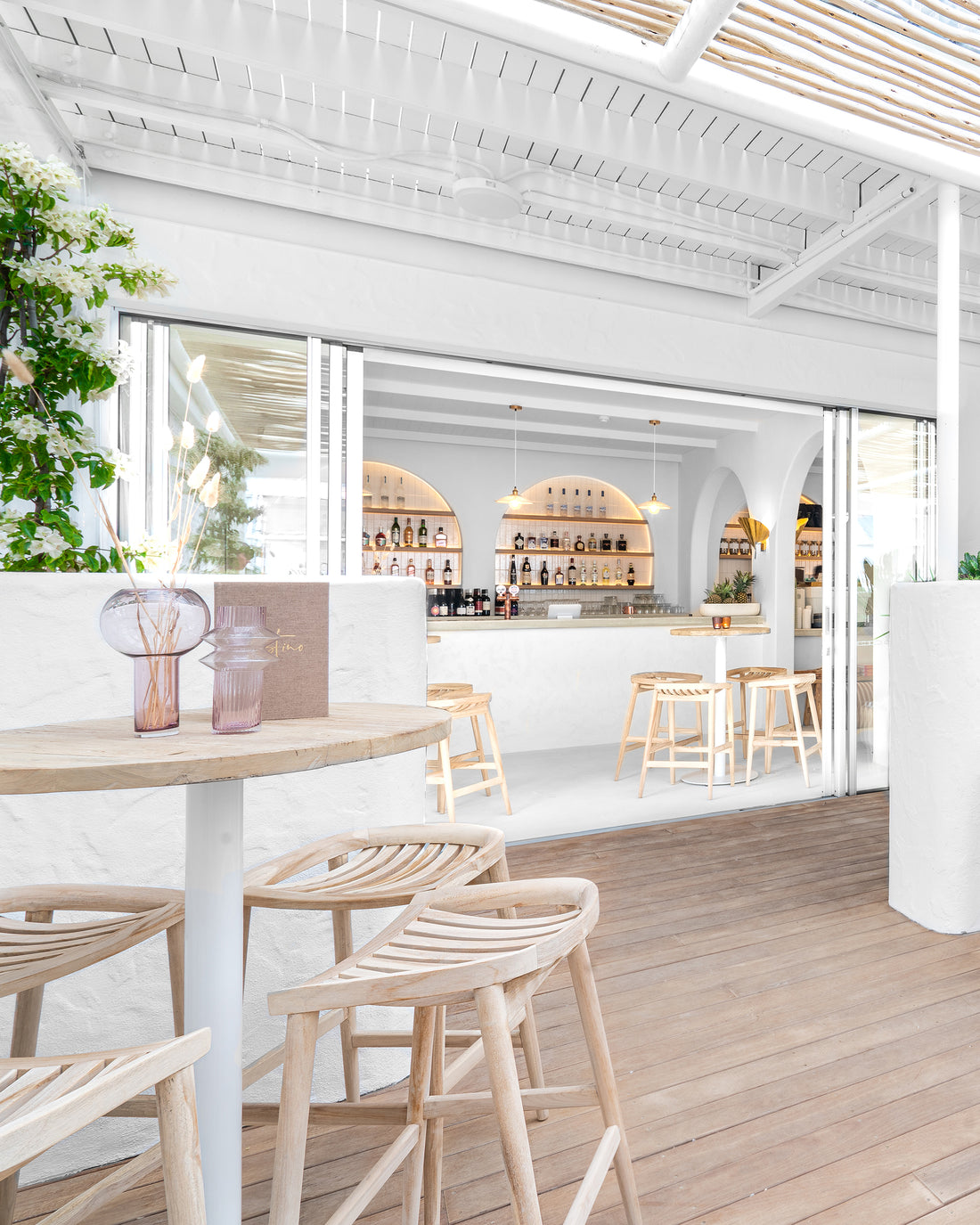 Uniqwa bar table setting with white pedestal base and natural teak bar stools in a stylish resort-inspired commercial venue.