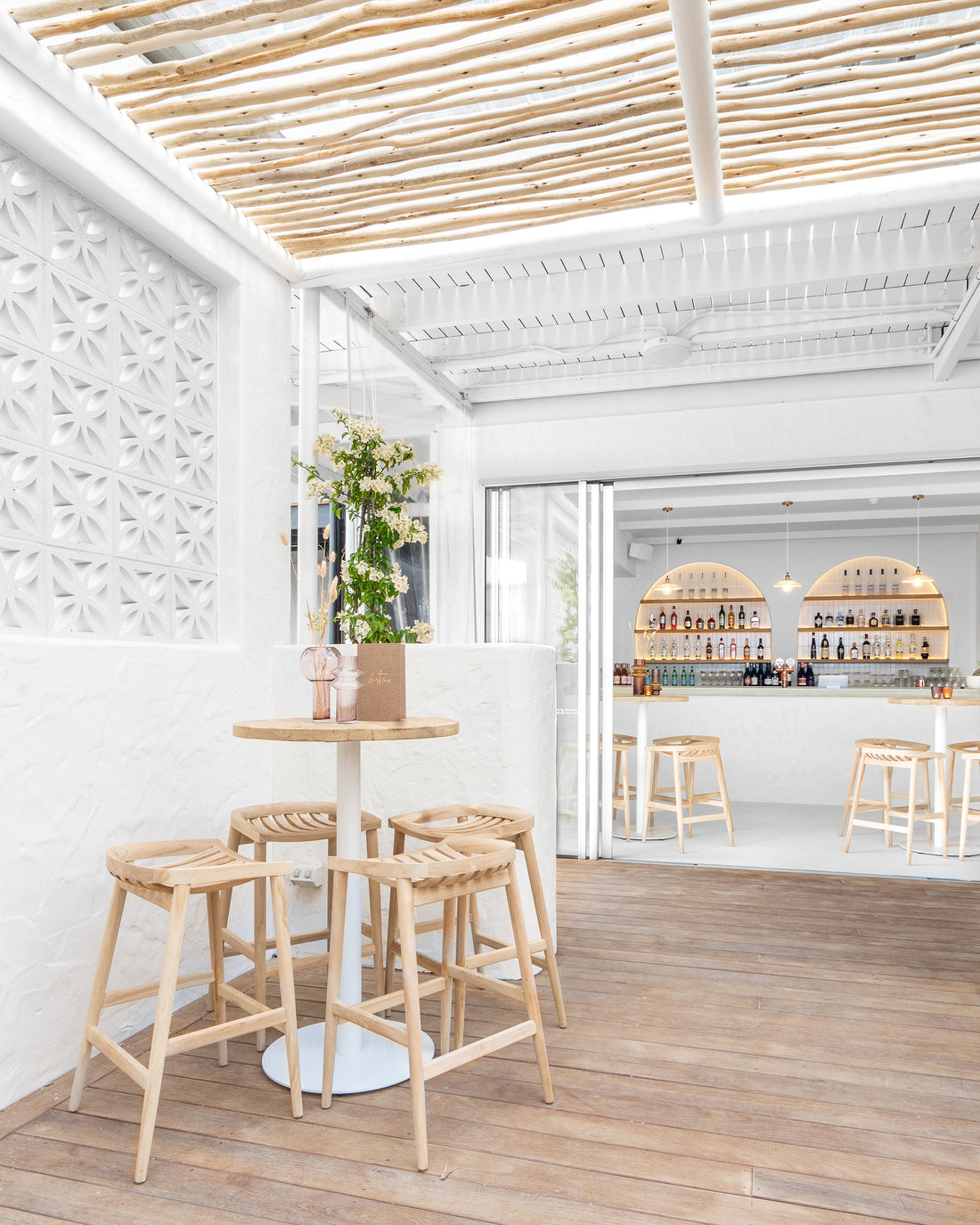 Uniqwa bar table setting with white pedestal base and natural teak bar stools in a stylish resort-inspired commercial venue.