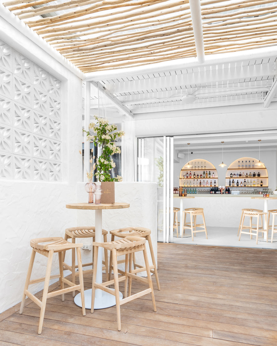 Uniqwa bar table setting with white pedestal base and natural teak bar stools in a stylish resort-inspired commercial venue.